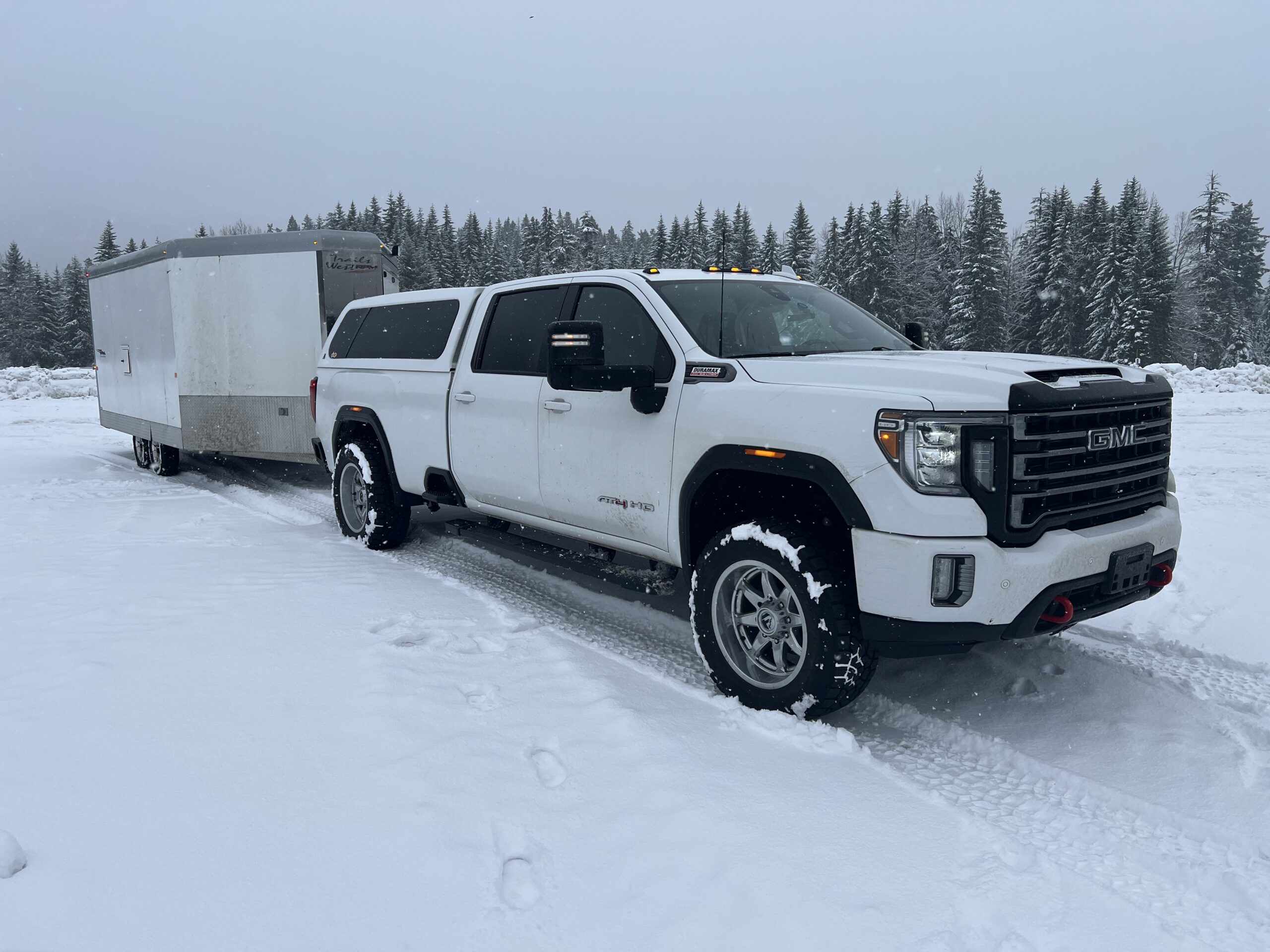 A white pickup truck with covered bed tows a trailer through the snow